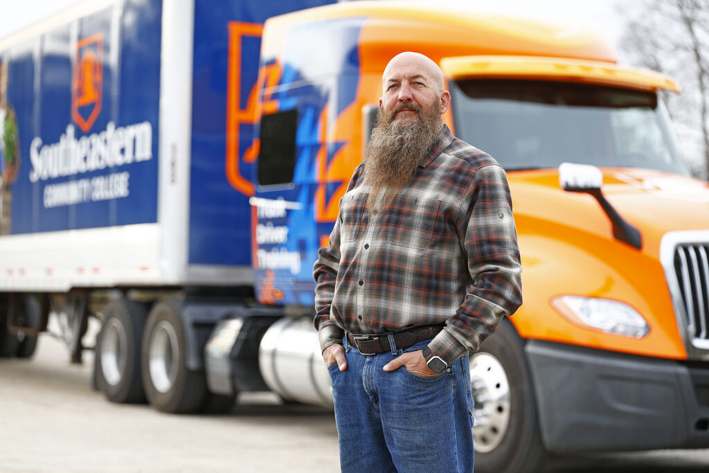 Truck Driver Training Instructor Will McCulloch stands in front of the new orange wrapped truck at the driving pad in Fair Bluff.