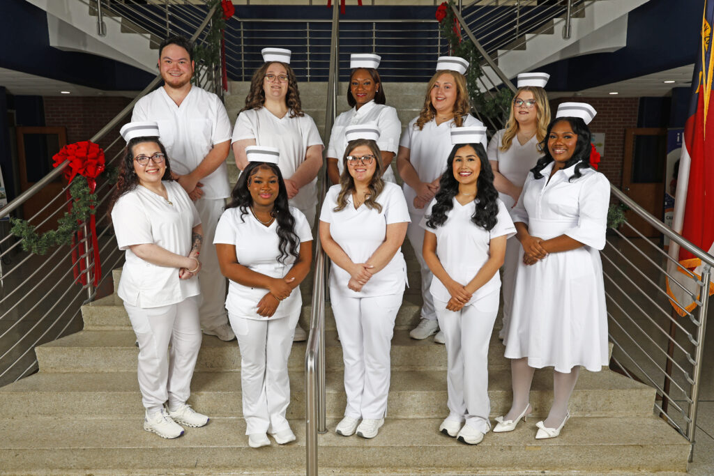 A group of practical nursing students in white uniforms pose together on a staircase inside a campus building. Students are wearing traditional nursing caps, and holiday garland decorates the stair railings behind them.