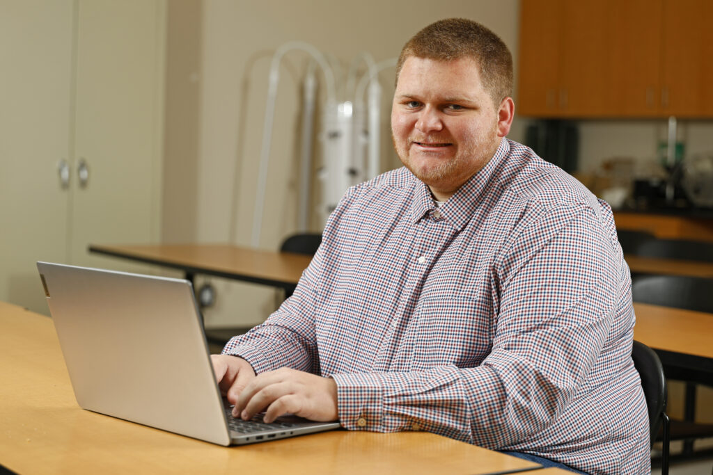Samuel Sibbett sitting at a desk in the Cartrette Building smiling at the camera while typing on a Laptop.