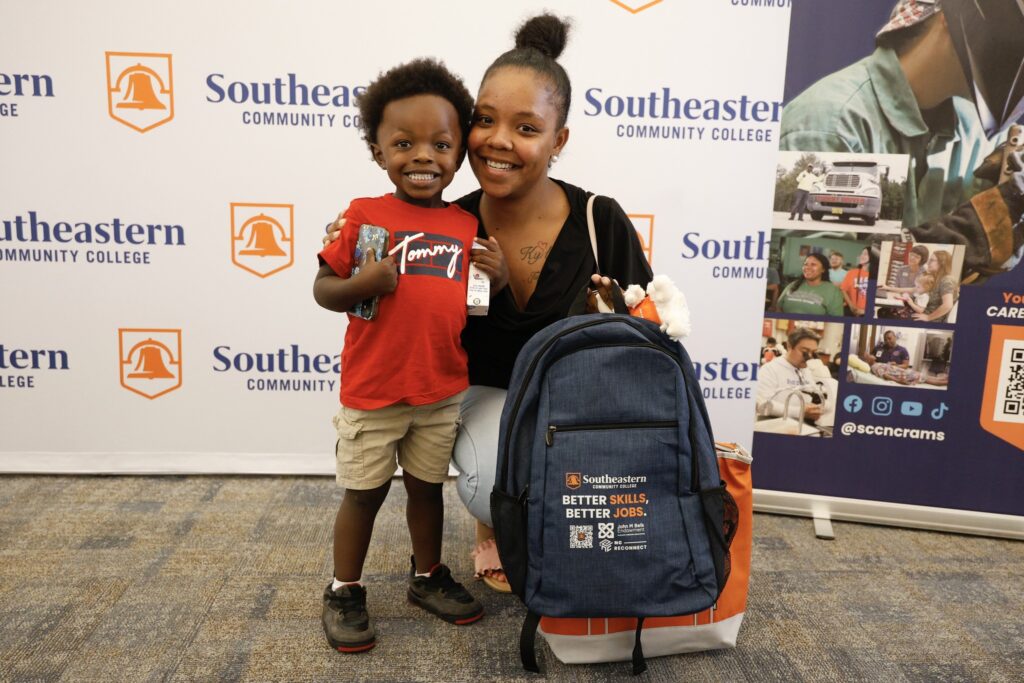 Student with her child at an NCReconnect event. Smiling at camera with a Better Skills, Better Jobs backpack.